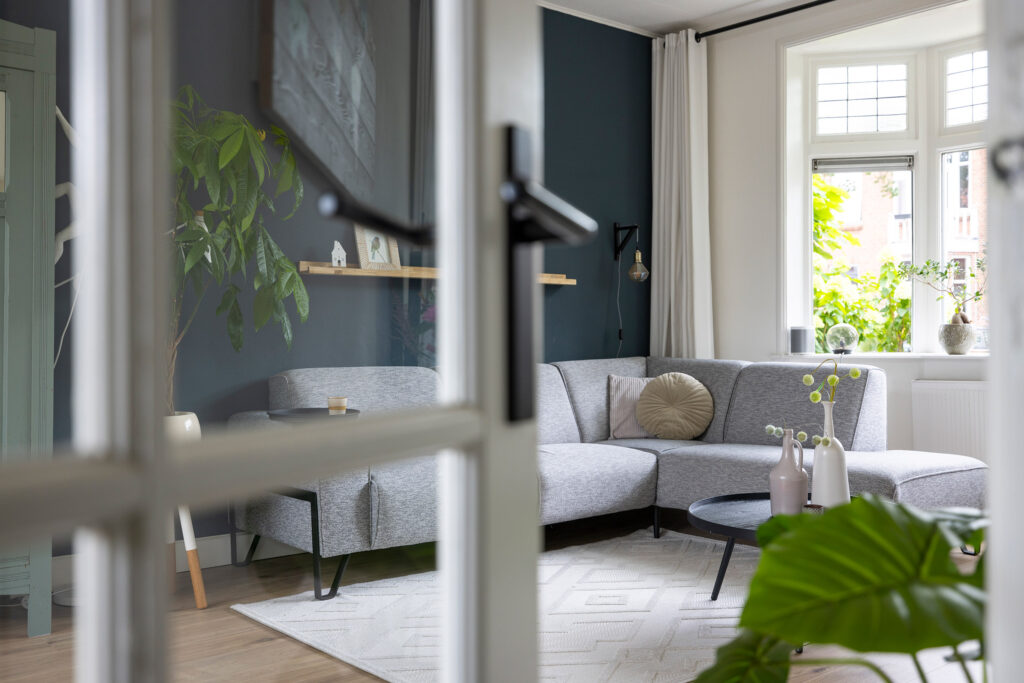 Woonkamer met grijze hoekbank, donkere wand en groot raam, gefotografeerd door glazen deuren