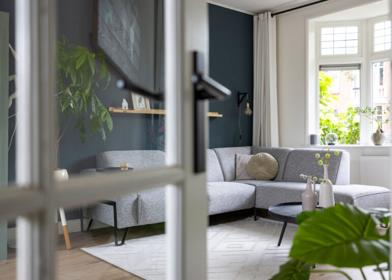 Woonkamer met grijze hoekbank, donkere wand en groot raam, gefotografeerd door glazen deuren