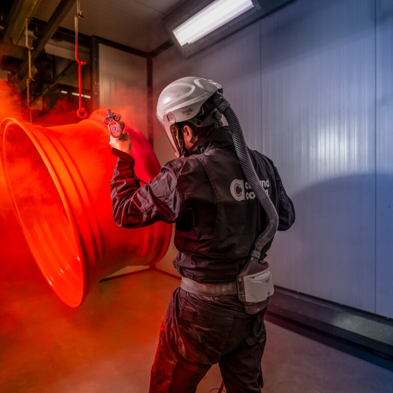 Medewerker met beschermende kap brengt een rode coating aan op hangende metalen velgen