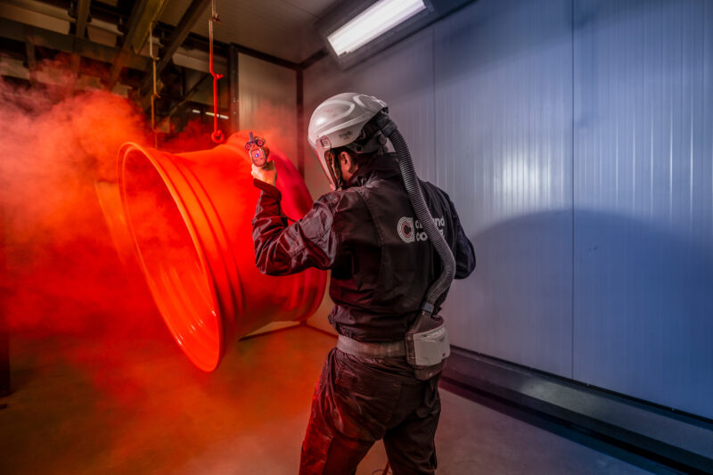 Medewerker met beschermende kap brengt een rode coating aan op hangende metalen velgen