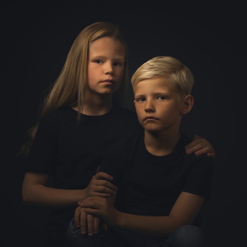 Zus met jonger broertje zittend voor haar met haar handen op zijn schouder voor een donkere achtergrond