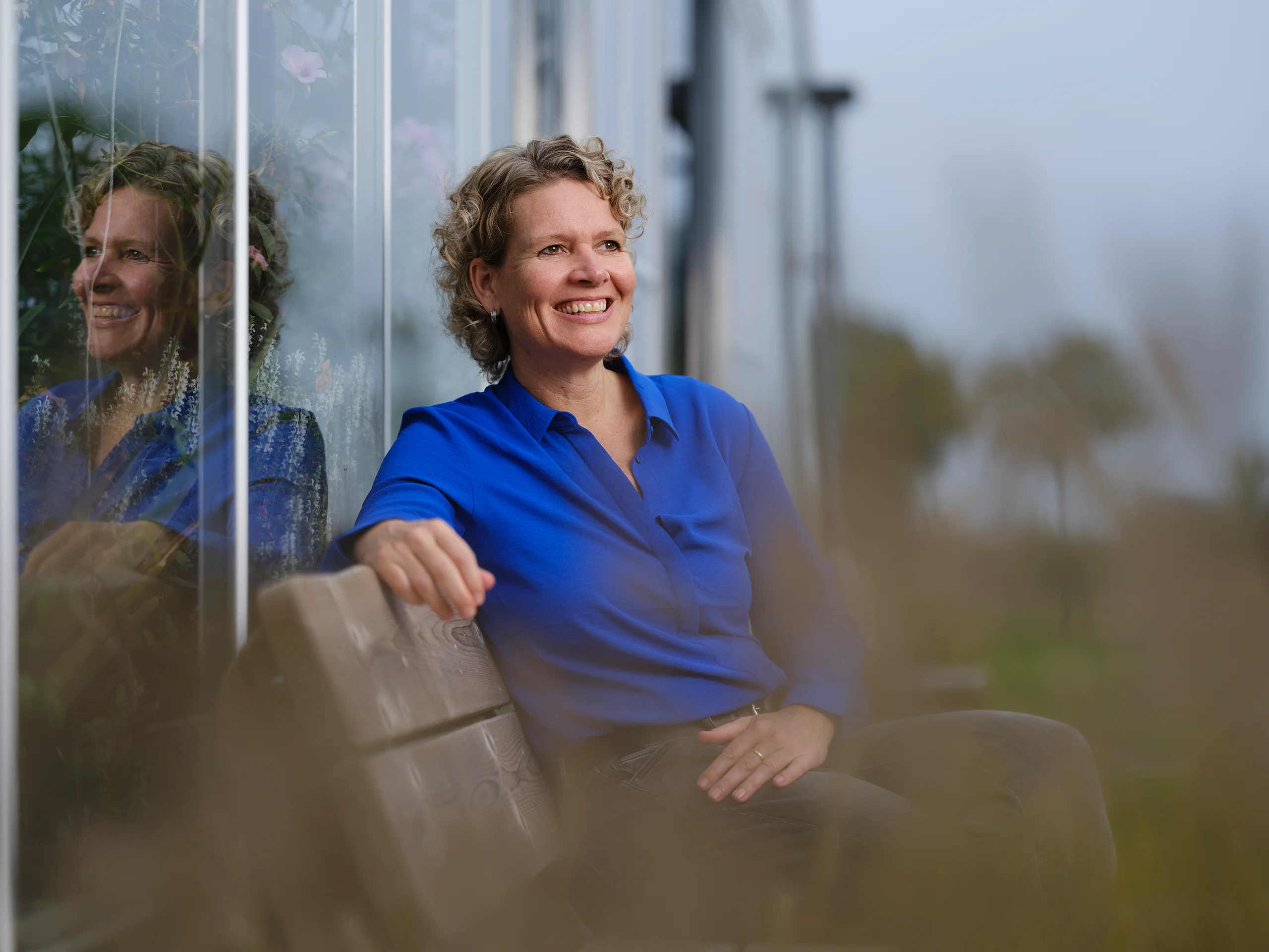 Zakelijk portret van blonde dame met krullen van middelbare leeftijd zittend op een bank met haar eigen weerspiegeling in het glas achter haar