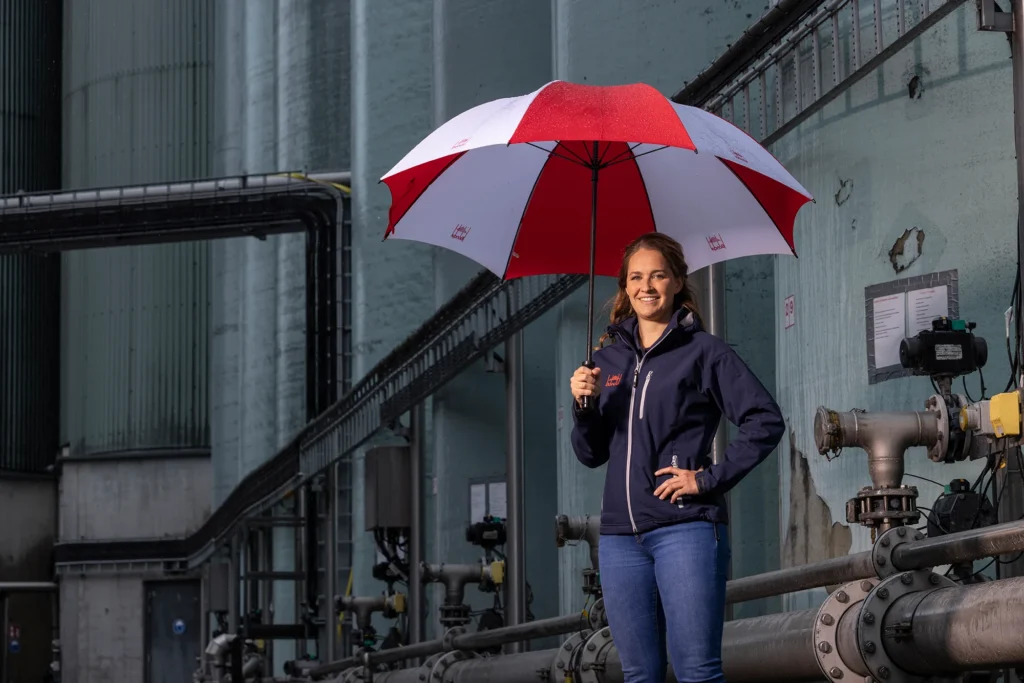 Dame met bruin haar, gekleed in blauwe spijkerbroek houdt een rood-witte paraplu vast en lacht richting de camera