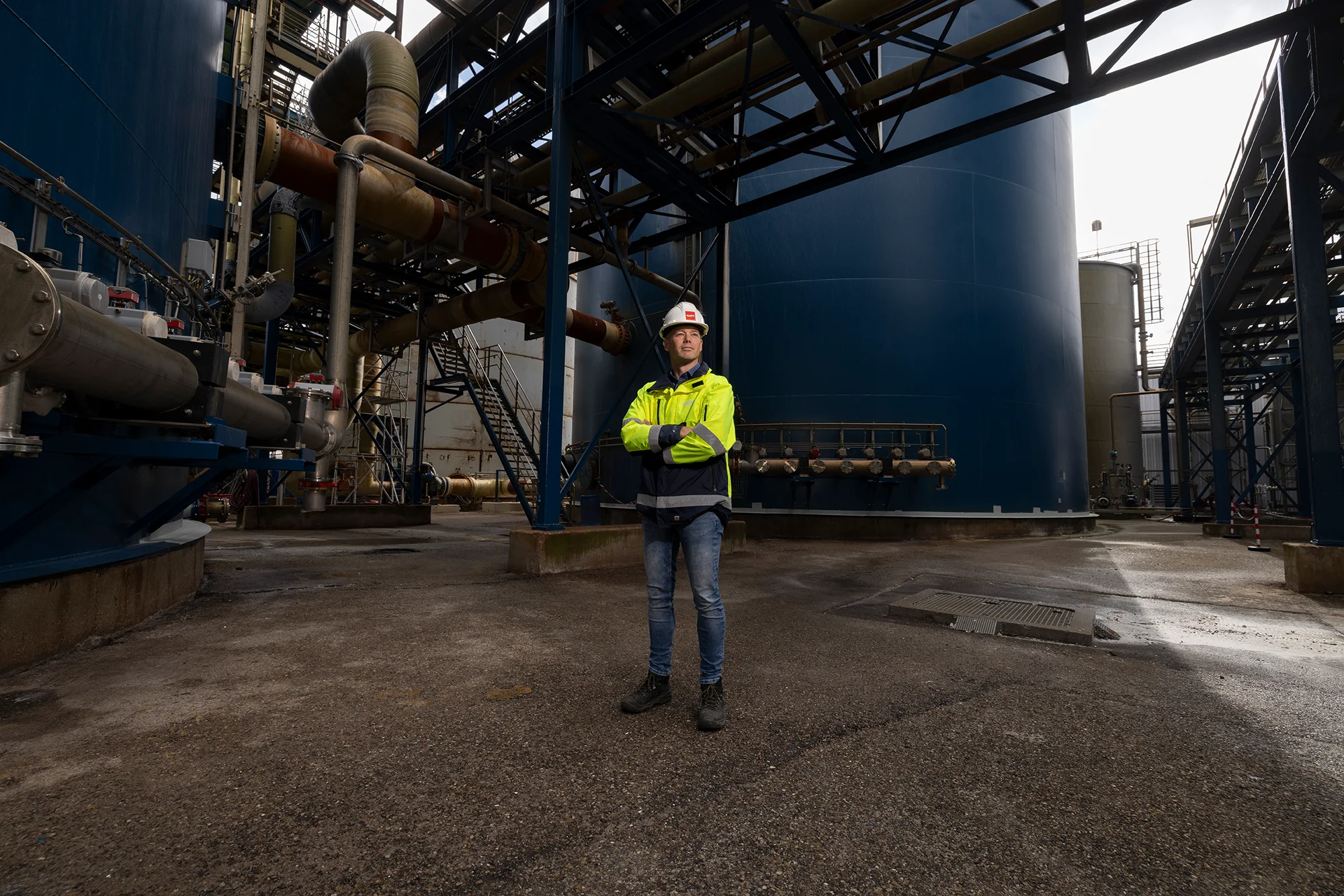 Projectmanager met veiligheidshelm en reflecterende jas tussen industriële installaties.
