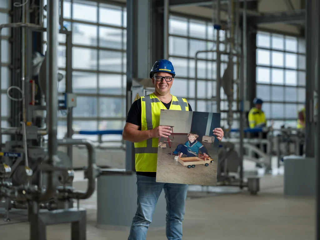 Ausnutria-medewerker met veiligheidshelm en reflecterend vest houdt zijn jeugdfoto vast in de fabriekshal.
