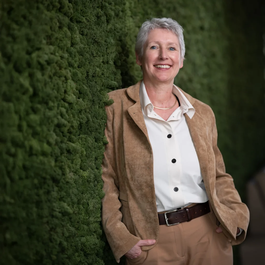 Dame in bruin tweedelig pak leunend tegen begroeide wand