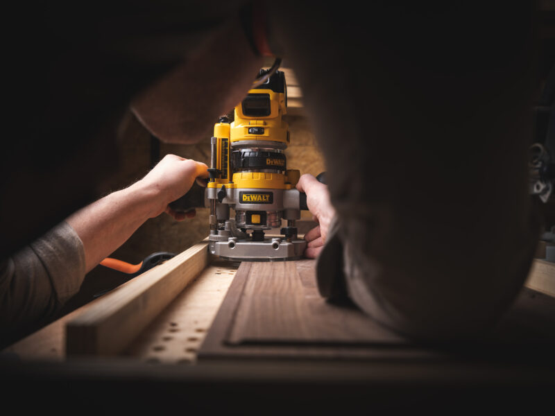 Houtbewerker gebruikt een gele bovenfrees aan een houten werkstuk in een sfeervol verlichte werkplaats.