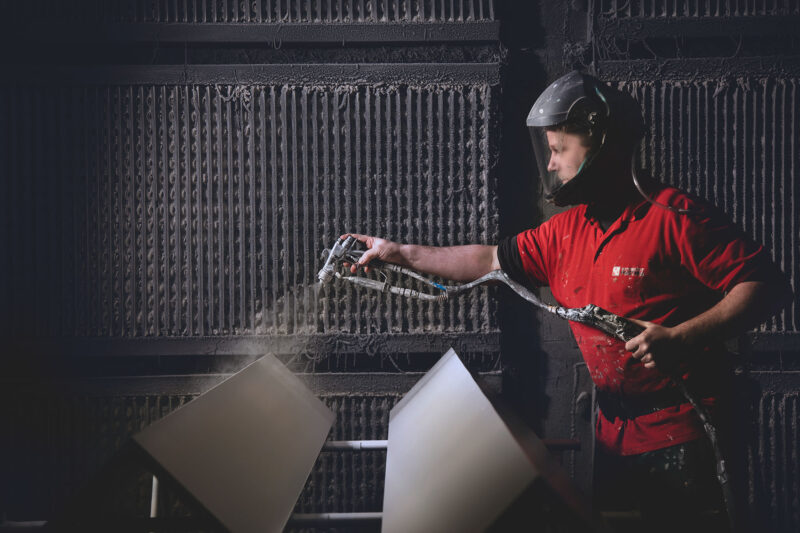 Medewerker met gelaatsbescherming spuit hangende metalen panelen in een spuitcabine