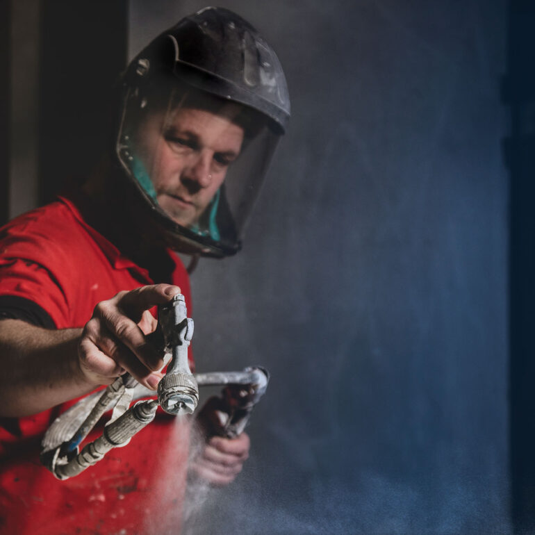 Vakman in een rood shirt en met gelaatsbescherming richt een spuitpistool op een metalen onderdeel