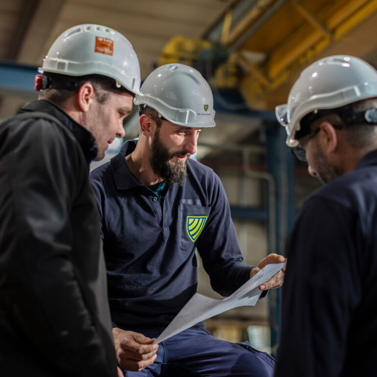 Drie medewerkers met veiligheidshelmen bespreken een werktekening in een productiehal