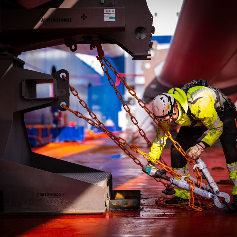 Medewerker met veiligheidshelm zet een grote lading vast met kettingen op het scheepsdek