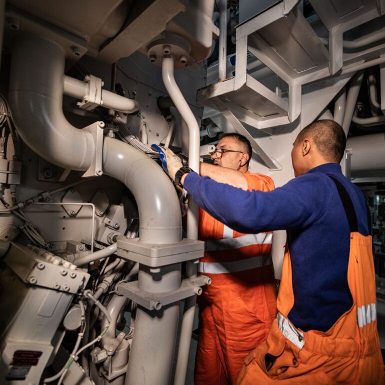 Twee medewerkers in oranje werkkleding werken aan leidingen en machines in een scheepsmachinekamer