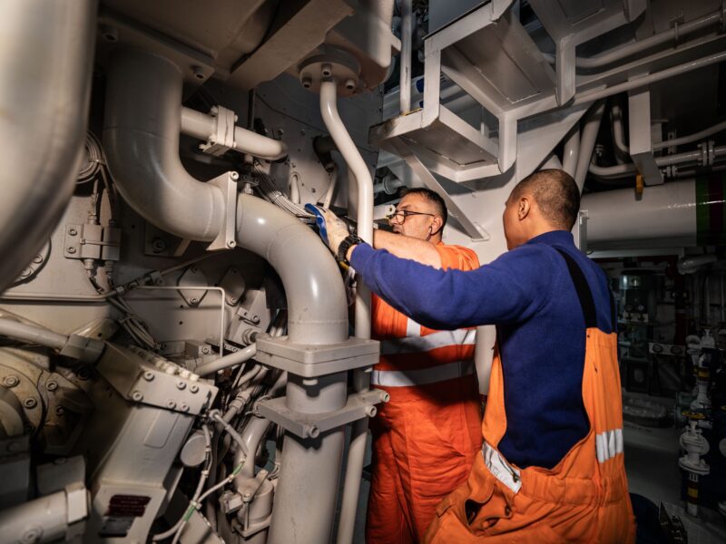 Twee medewerkers in oranje werkkleding werken aan leidingen en machines in een scheepsmachinekamer