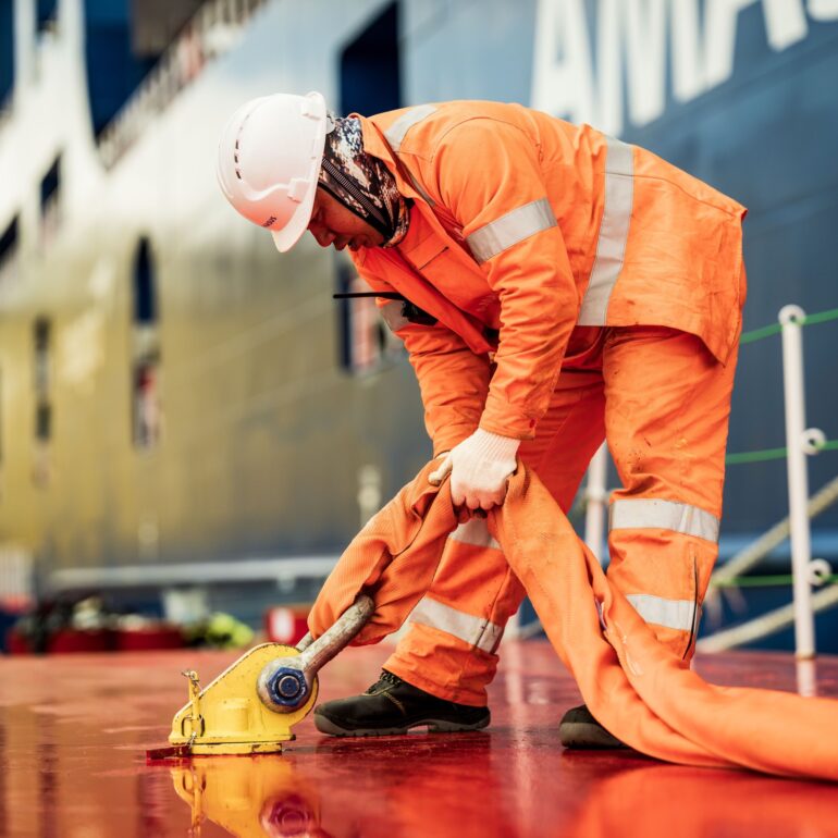 Medewerker in oranje werkkleding spant een brede band met een ratel op het scheepsdek