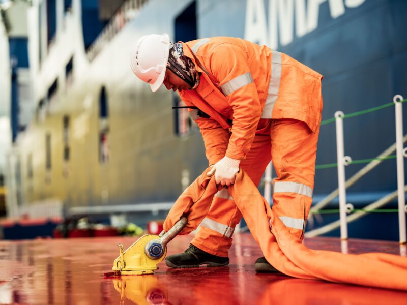 Medewerker in oranje werkkleding spant een brede band met een ratel op het scheepsdek