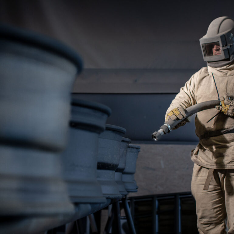 Straler met beschermende helm behandelt grote metalen onderdelen in een straalcabine