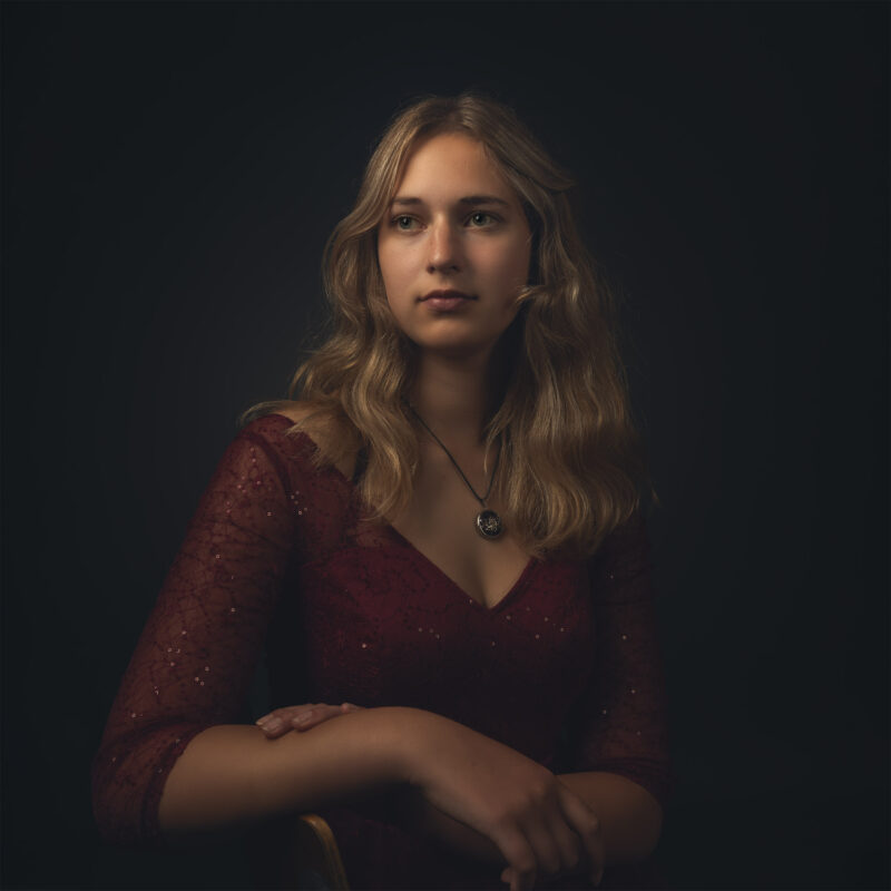 Portret van een vrouw met lang haar in een rode jurk, zittend op een stoel.