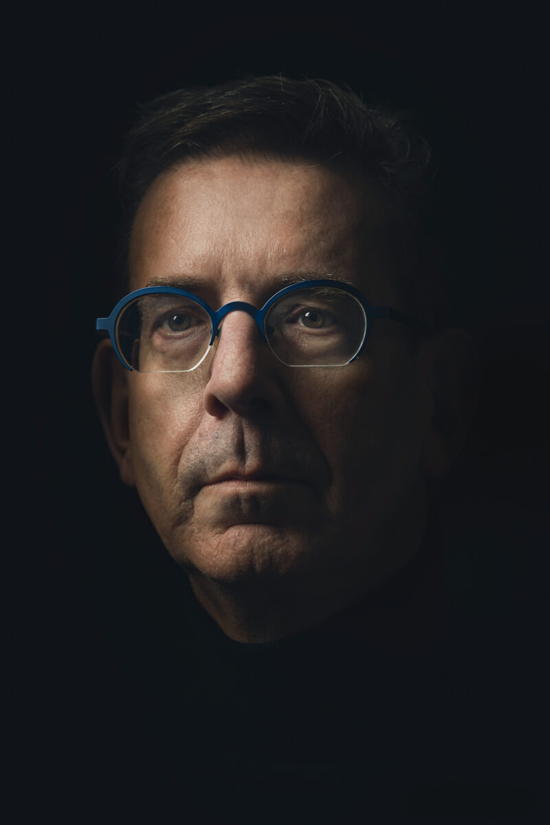 Portret van een man met een bril, grijs haar en een snor tegen een donkere achtergrond.