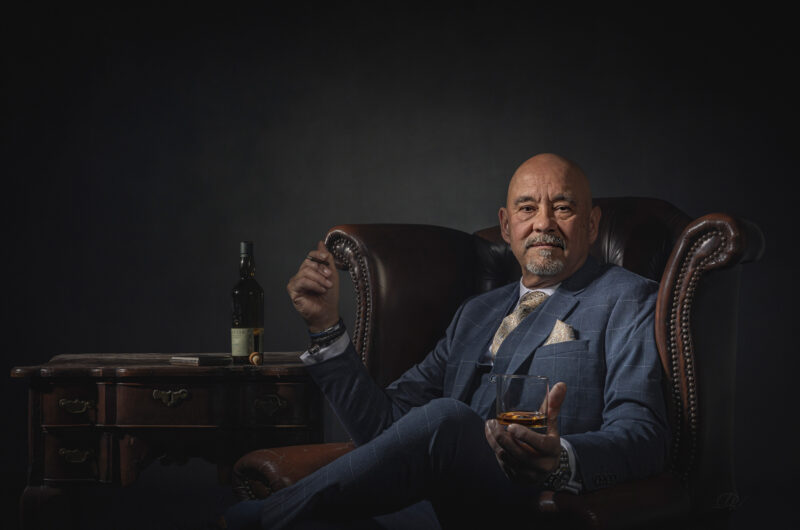Geënsceneerd portret van een man in een pak, zittend in een stoel met een glas.