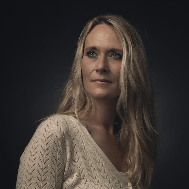 Vrouw met lang blond haar en een lichte gebreide trui tegen een donkere achtergrond.