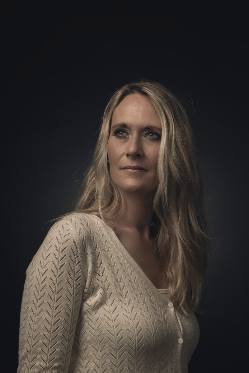Portret van een vrouw met lang blond haar en een lichte trui tegen een donkere achtergrond.