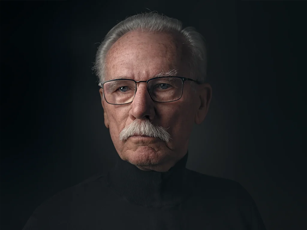 Portret van een man met grijs haar, een snor en een bril tegen een donkere achtergrond.