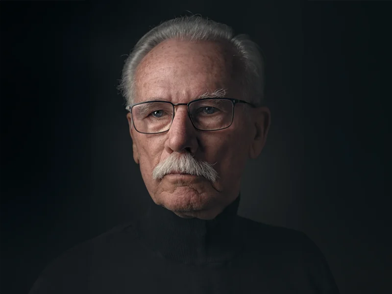 Portret van een man met grijs haar, een snor en een bril tegen een donkere achtergrond.
