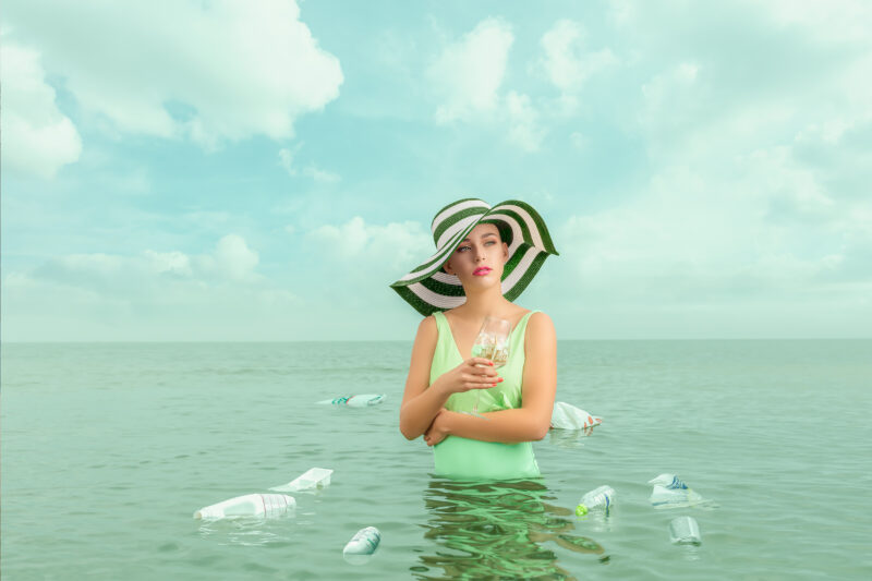 Vrouw met een groen-witte hoed staat in zee met een glas water, tussen drijvende plastic flessen.