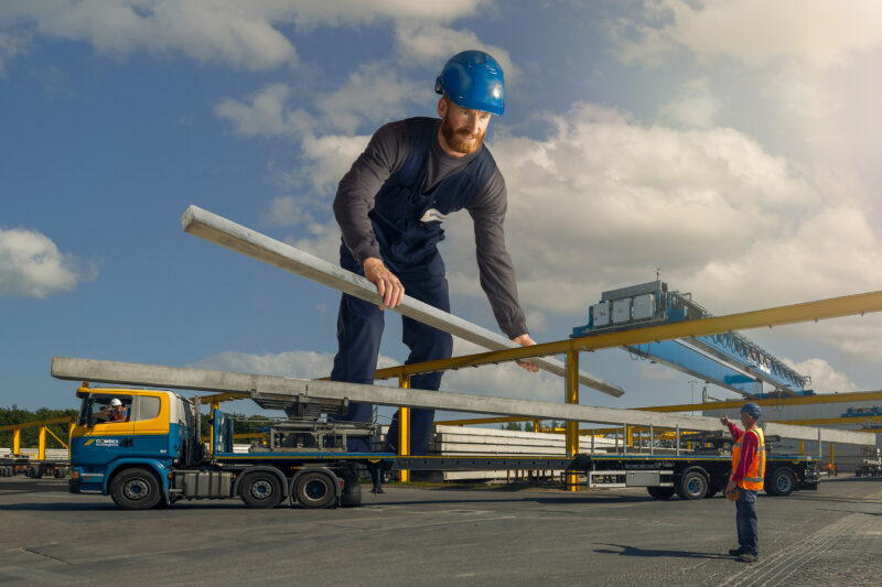 Reusachtige medewerker met een blauwe veiligheidshelm houdt een betonnen balk boven een vrachtwagen.