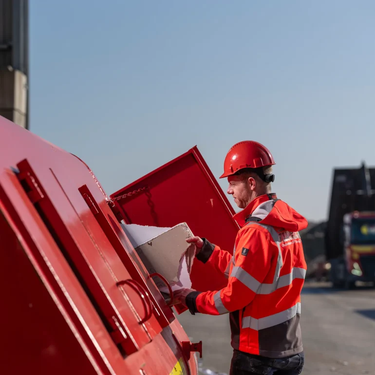Medewerker met rode veiligheidshelm en oranje werkkleding plaatst afval in een rode container op een bedrijfsterrein