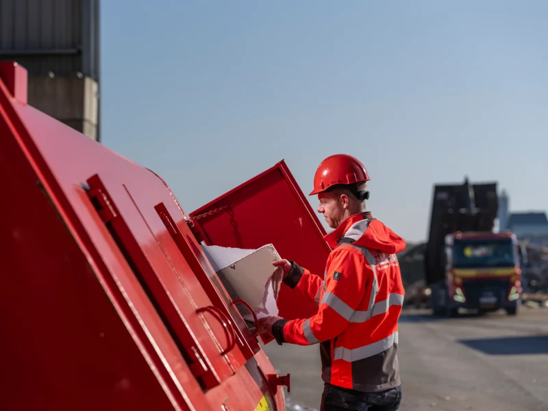 Medewerker met rode veiligheidshelm en oranje werkkleding plaatst afval in een rode container op een bedrijfsterrein