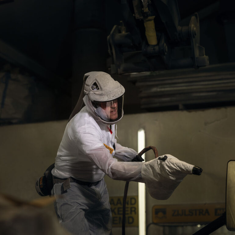 Medewerker in een witte beschermende overall houdt spuitapparatuur vast in de werkplaats