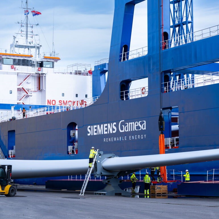 Medewerkers staan bij een lang windturbineonderdeel naast de blauwe constructie van een transportschip