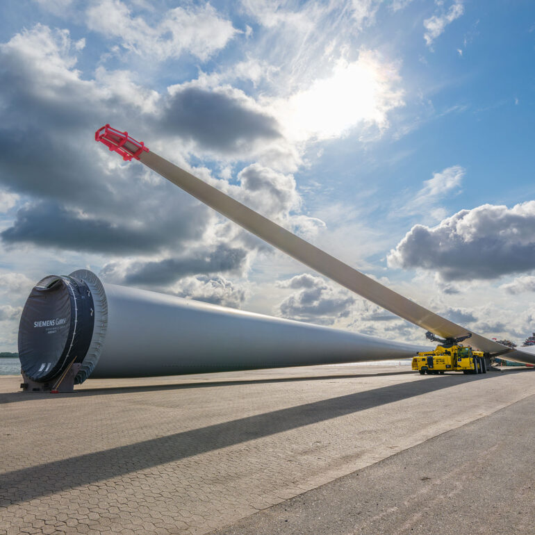 Lang windturbineblad op transportmaterieel op een haventerrein onder een bewolkte lucht