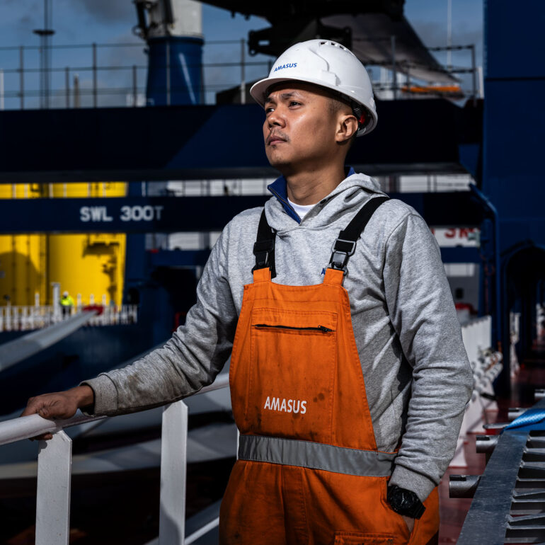 Medewerker van Amasus met witte veiligheidshelm en oranje overall staat bij de reling van een schip