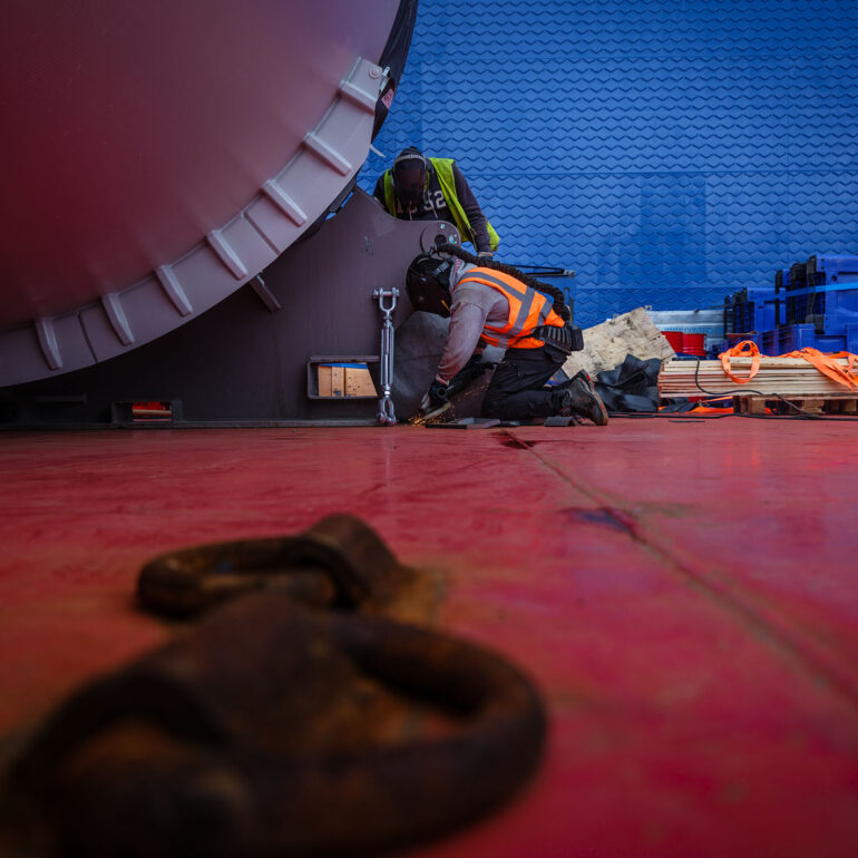 Twee medewerkers zetten een groot industrieel onderdeel vast op een rood scheepsdek