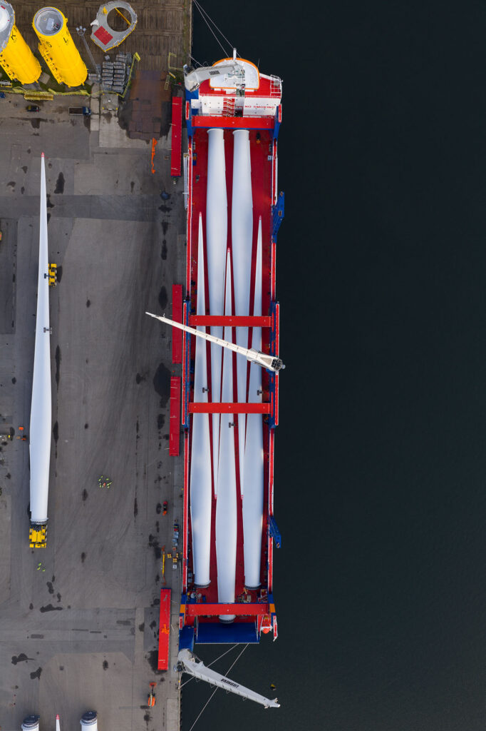 Bovenaanzicht van een vrachtschip met lange windturbinebladen aan een havenkade