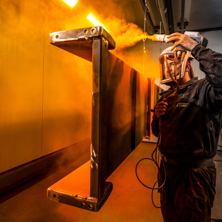 Medewerker met beschermende kap spuit een hangend metalen frame in warm verlichte nevel