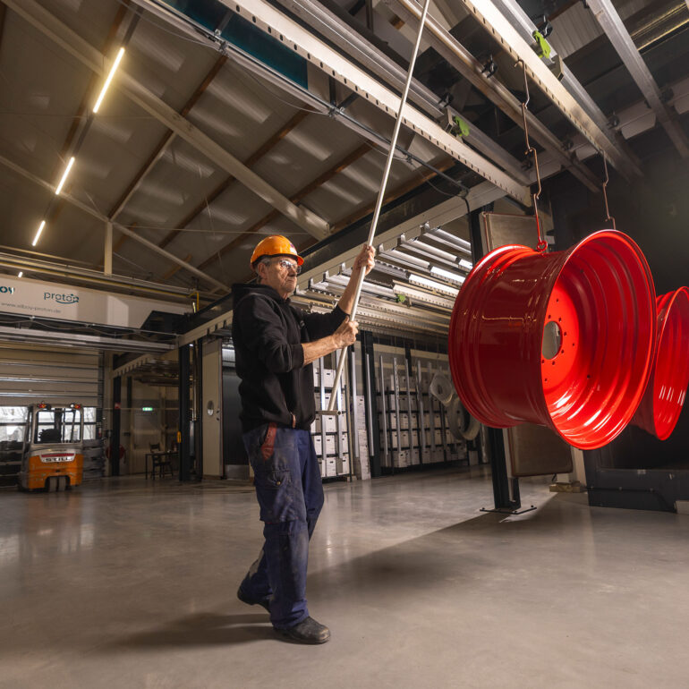 Medewerker met veiligheidshelm begeleidt rode metalen velgen aan een hangend transportsysteem