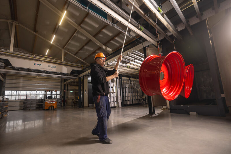 Medewerker met veiligheidshelm begeleidt rode metalen velgen aan een hangend transportsysteem