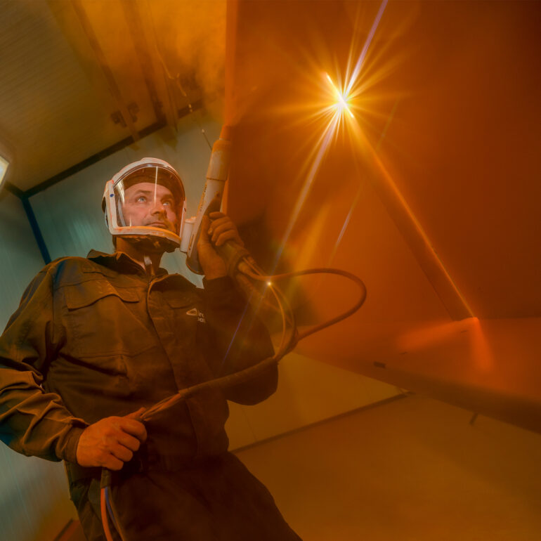 Vakman met beschermende kap houdt een spuitpistool vast in een spuitcabine met warm tegenlicht