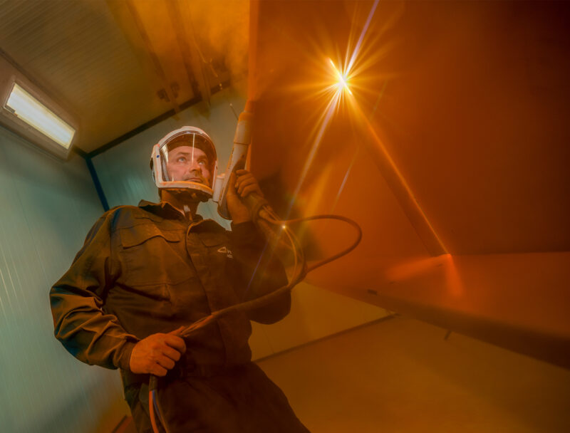 Vakman met beschermende kap houdt een spuitpistool vast in een spuitcabine met warm tegenlicht