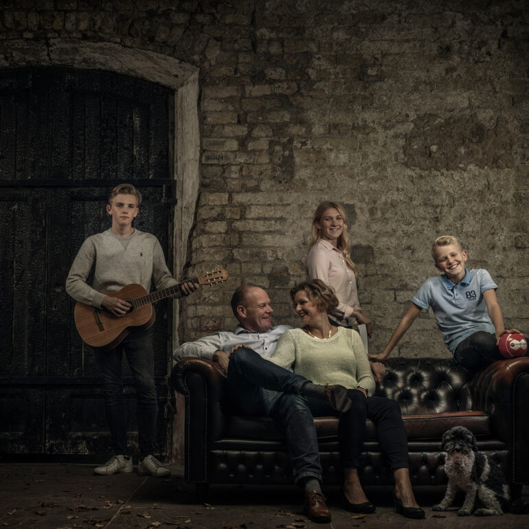 Gezin met drie kinderen poseert rond een leren bank bij oude stenen muren, met een gitaar, een bal en een hond.
