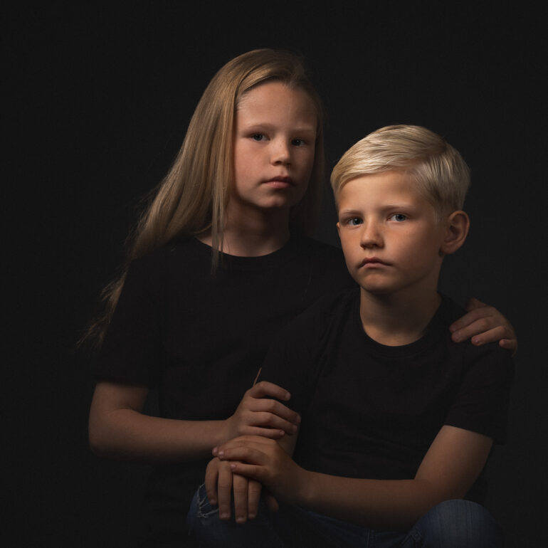 Jongen en meisje in donkere kleding zitten dicht bij elkaar tegen een donkere achtergrond.