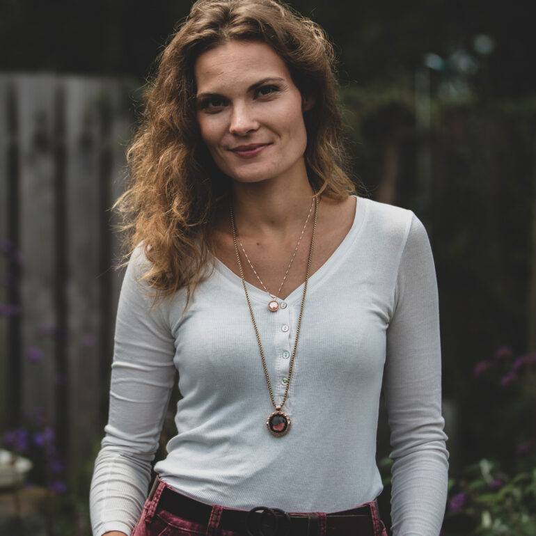 Vrouw met krullend haar, een lichte top en een lange ketting staat voor een groene, onscherpe achtergrond.