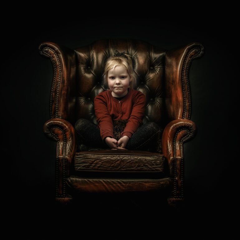 Jong kind in een rode trui zit op een grote donkerleren fauteuil tegen een zwarte achtergrond.