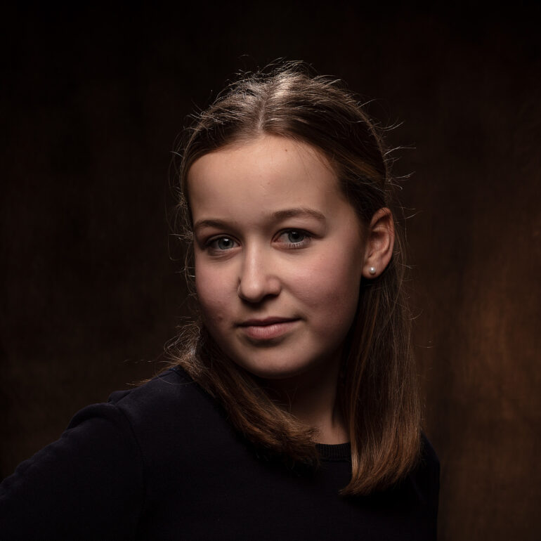 Jonge vrouw met halflang bruin haar en een donkere top kijkt naar de camera tegen een warme bruine achtergrond.