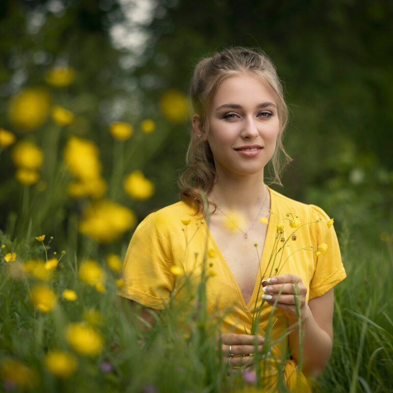 Jonge vrouw in een gele blouse staat tussen groen gras en gele bloemen.