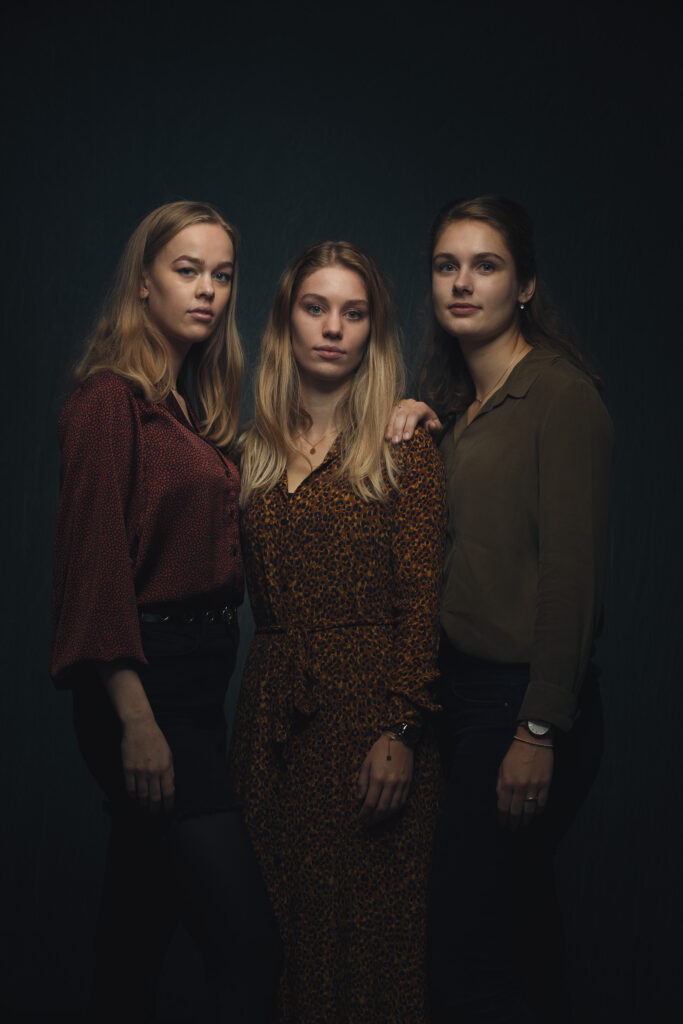Portret van drie zussen die naast elkaar staan tegen een donkere achtergrond.