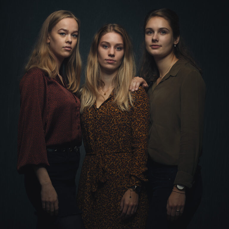 Drie jonge vrouwen staan dicht naast elkaar tegen een donkere achtergrond.